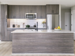 Studios in Arlington, VA - Sage at National Landing - Kitchen with Stainless Steel Appliances, White Countertops, White Tile Backsplash, Wood-Style Cabinets, Wood-Style Flooring, a Large Window, and a Glassdoor